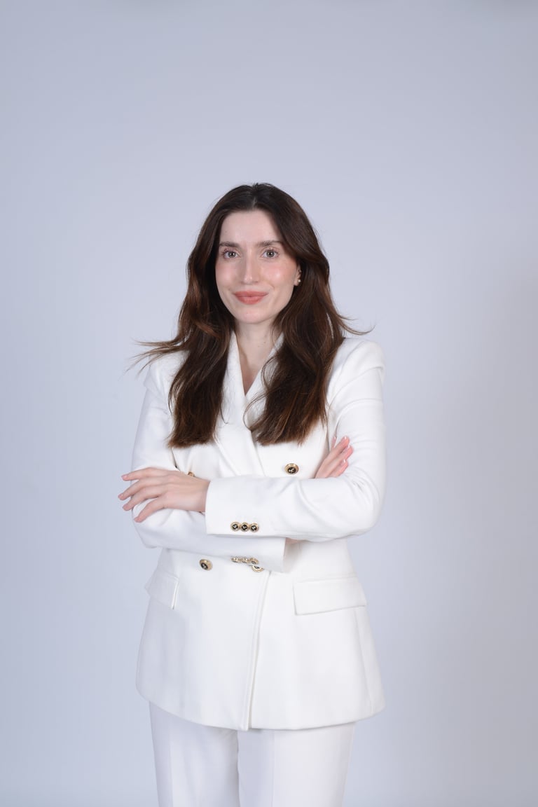 Professional headshot of a woman with long dark hair wearing a white blazer and pants against a light gray background
