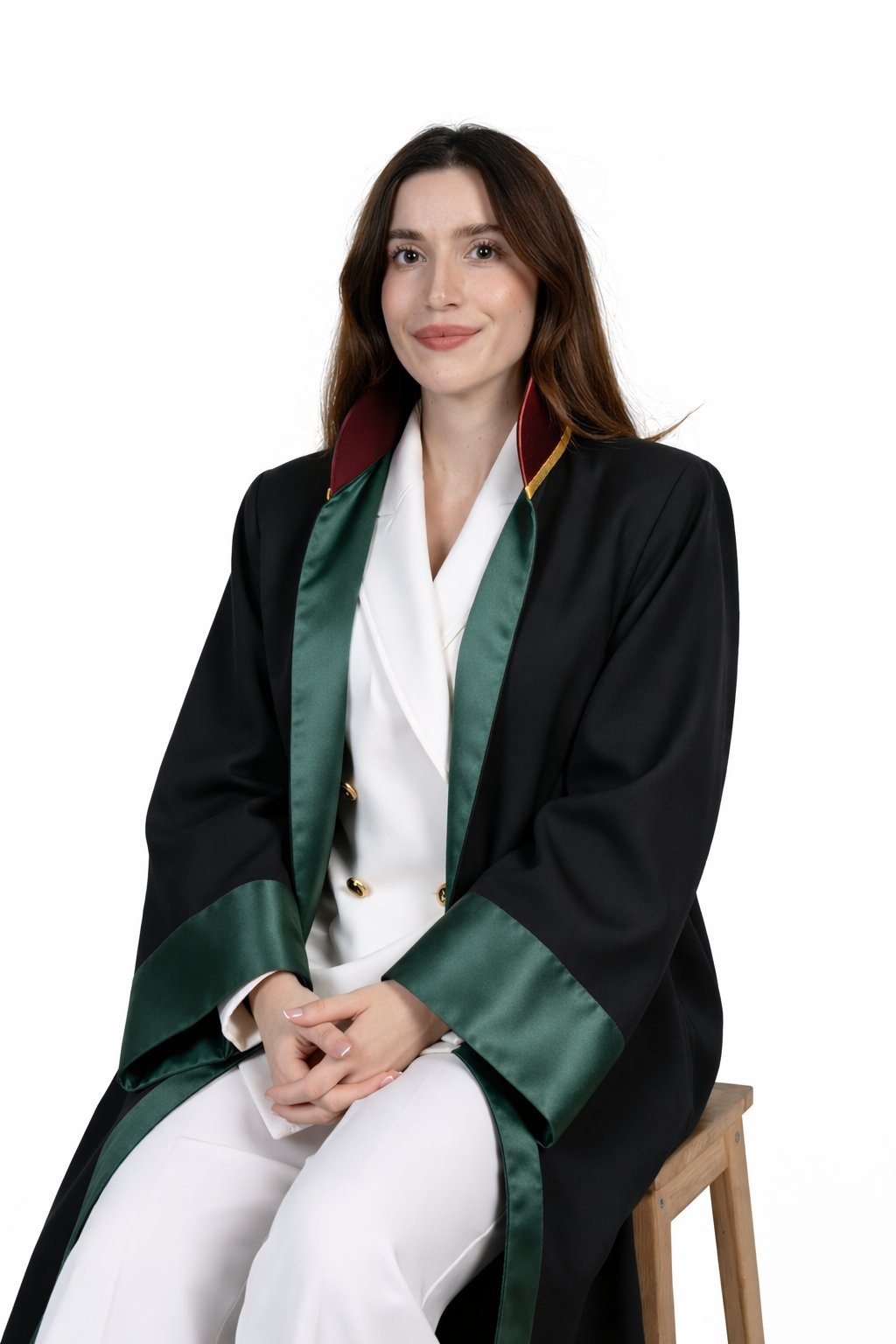 Woman sitting on wooden stool wearing black blazer with green satin lapels over white blouse, against white background